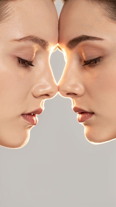 Two women share a peaceful moment with foreheads gently touching in this minimalist portrait featuring soft pearl-gray tones and elegant composition.