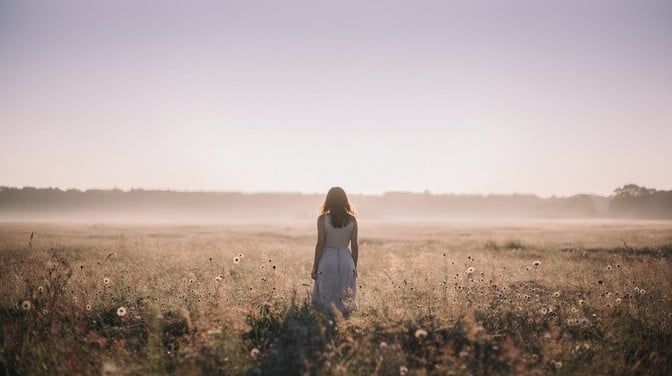 A young woman stands alone in a misty meadow at golden hour, surrounded by wildflowers and soft pastel skies creating a dreamlike scene.
